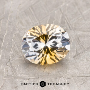 A 1.39-carat yellow-colorless particolored Montana Sapphire (heated) is displayed on a textured gray surface, with "EARTH'S TREASURY" text and logo below.