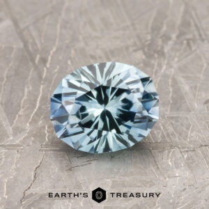 A 1.29-carat heated aqua blue Montana Sapphire, cut in a round brilliant style, sits on a textured metallic surface with the text "EARTH'S TREASURY" below.