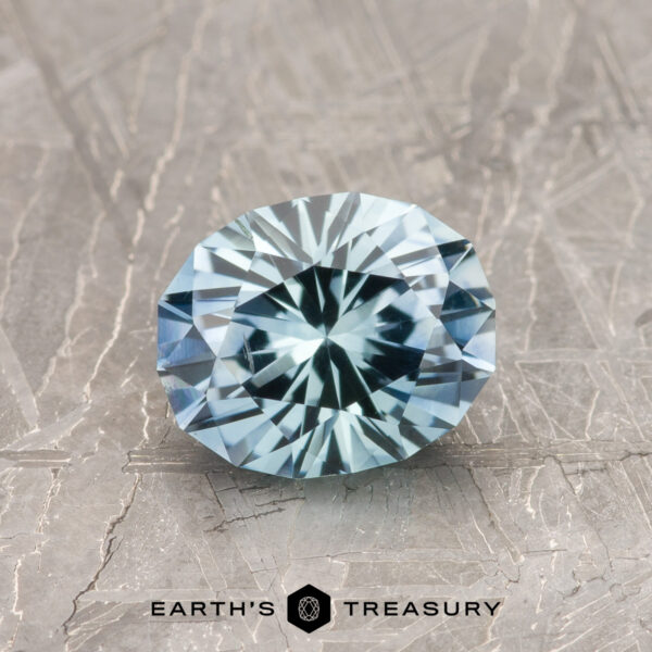 1.29-Carat Aqua Blue Montana Sapphire (Heated) A 1.29-carat heated aqua blue Montana Sapphire, cut in a round brilliant style, sits on a textured metallic surface with the text "EARTH'S TREASURY" below.
