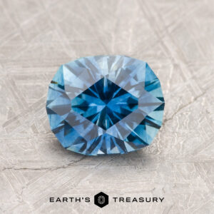A 1.67-carat medium blue Montana Sapphire (heated) is displayed on a textured gray surface with the text "Earth's Treasury" and a hexagon logo beneath it.