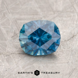 A 1.36-carat heated teal blue Montana Sapphire rests on a textured gray surface, with "Earth's Treasury" below it.