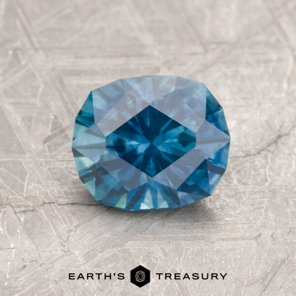 1.36-Carat Teal Blue Montana Sapphire (Heated) A 1.36-carat heated teal blue Montana Sapphire rests on a textured gray surface, with "Earth's Treasury" below it.