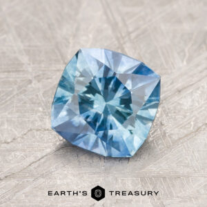 A 1.25-carat blue-aqua particolored heated Montana Sapphire with cushion cut is showcased on a textured light surface. The "Earth's Treasury" logo is displayed at the bottom.