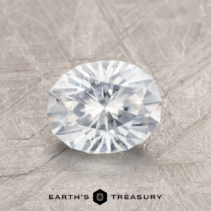 A 1.41-Carat Colorless Montana Sapphire with a round faceted cut rests on a textured gray surface, with the Earth's Treasury logo and text below.