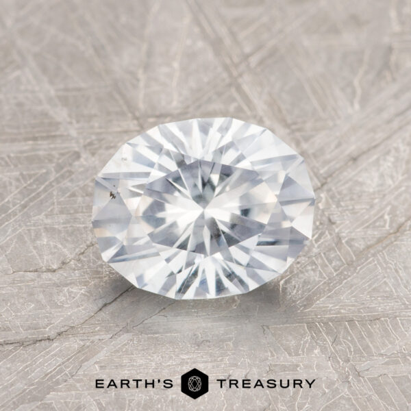1.41-Carat Colorless Montana Sapphire A 1.41-Carat Colorless Montana Sapphire with a round faceted cut rests on a textured gray surface, with the Earth's Treasury logo and text below.