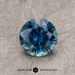 A 1.39-carat rich teal blue Montana Sapphire (heated), round-cut, is displayed on a textured gray surface with "EARTH'S TREASURY" and a geometric logo at the bottom.