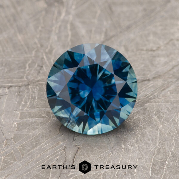 1.39-Carat Rich Teal Blue Montana Sapphire (Heated) A 1.39-carat rich teal blue Montana Sapphire (heated), round-cut, is displayed on a textured gray surface with "EARTH'S TREASURY" and a geometric logo at the bottom.