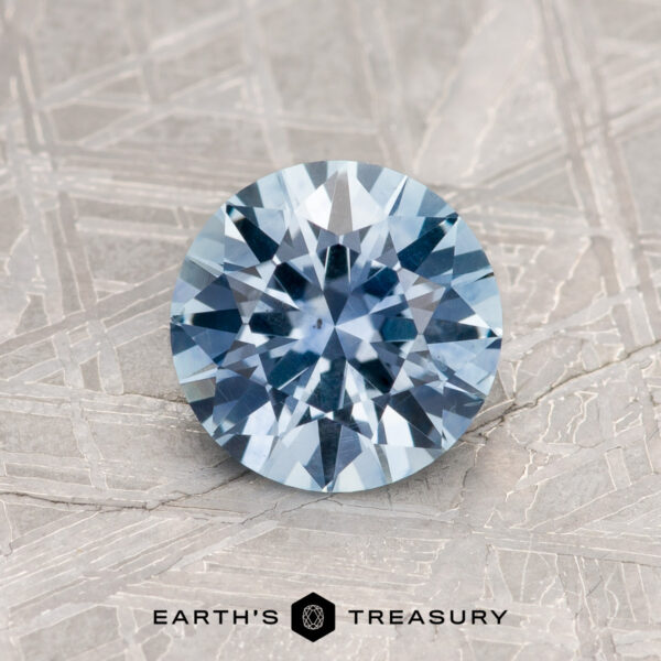 1.31-Carat Light Sky Blue Montana Sapphire (Heated) A 1.31-carat light sky blue Montana sapphire (heated), round brilliant-cut, is displayed on a textured metallic surface with the text "EARTH'S TREASURY" visible below.