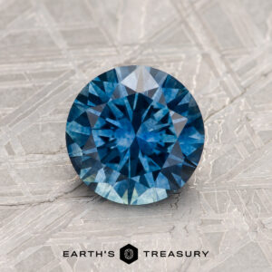 A 1.30-Carat Peacock Blue Montana Sapphire (Heated), round brilliant cut, sits on a patterned metallic surface with "EARTH'S TREASURY" text below.