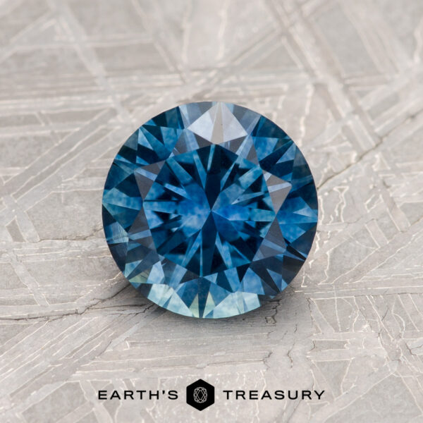 1.30-Carat Peacock Blue Montana Sapphire (Heated) A 1.30-Carat Peacock Blue Montana Sapphire (Heated), round brilliant cut, sits on a patterned metallic surface with "EARTH'S TREASURY" text below.