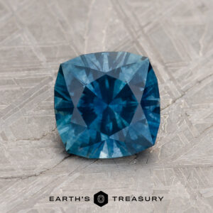 A 1.44-Carat Deep Peacock Blue Montana Sapphire (Heated), cushion-cut, rests on a textured gray surface with the "Earth's Treasury" logo and text below.