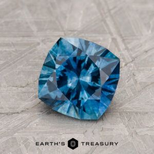 A 1.36-carat heated Peacock Blue Montana Sapphire is shown cushion-cut on a textured gray surface, with "EARTH'S TREASURY" and its logo featured at the bottom.