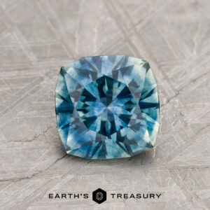 A 1.22-carat Teal Blue Montana Sapphire (Heated), cushion-cut, is shown on a textured gray surface with the "EARTH'S TREASURY" text and a geometric logo beneath it.