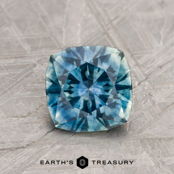 1.22-Carat Teal Blue Montana Sapphire (Heated) A 1.22-carat Teal Blue Montana Sapphire (Heated), cushion-cut, is shown on a textured gray surface with the "EARTH'S TREASURY" text and a geometric logo beneath it.