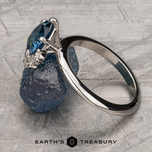 The “Alani” ring in 14k white gold with 1.28-Carat Montana Sapphire The “Alani” ring in 14k white gold, featuring a 1.28-carat Montana Sapphire, rests against a rough blue crystal on a textured gray surface. "EARTH'S TREASURY" is displayed at the bottom.