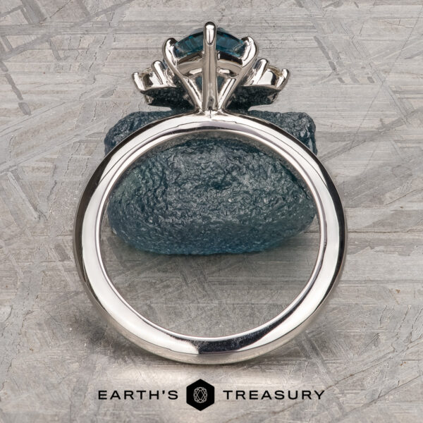 The “Alani” ring in 14k white gold with 1.28-Carat Montana Sapphire The Stacked from 55 images. Method=B (R=1,S=1) ring with a large blue gemstone is displayed upright against a rough blue stone on a textured gray surface, with the Earth’s Treasury logo at the bottom.