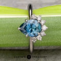 The “Alani” ring in 14k white gold with a 1.28-carat Montana sapphire is displayed on a green background, accented by six small round diamonds and the "Earth's Treasury" jewelry logo below.