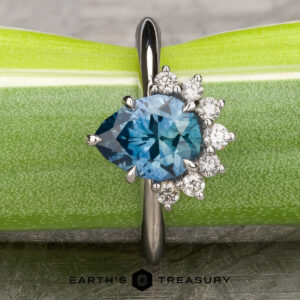 The “Alani” ring in 14k white gold with a 1.28-carat Montana sapphire is displayed on a green background, accented by six small round diamonds and the "Earth's Treasury" jewelry logo below.