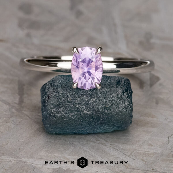 The "Fleur" ring in 14K white gold with 1.06-carat Umba sapphire The "Fleur" ring in 14K white gold, featuring a 1.06-carat faceted Umba sapphire, is displayed upright on a blue-green stone, with the "Earth's Treasury" logo and text below.