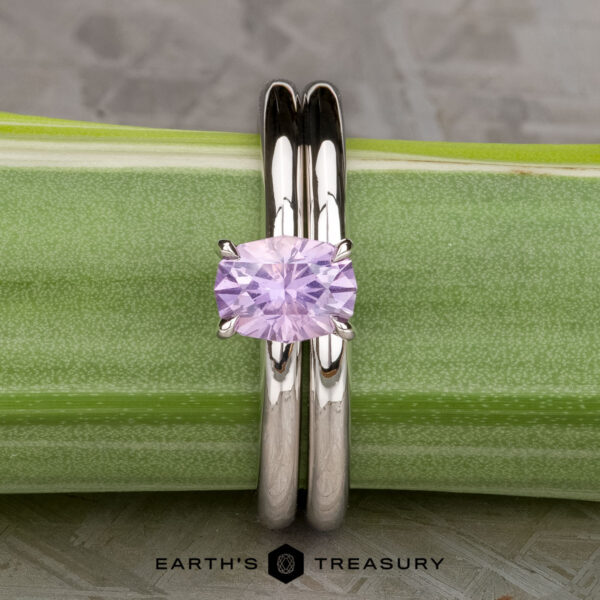 The "Fleur" ring in 14K white gold with 1.06-carat Umba sapphire, alongside the custom-fit straight band in 14K white gold The "Fleur" ring in 14K white gold with a 1.06-carat Umba sapphire is shown upright next to a green plant stem, alongside its custom-fit straight band; the "Earth's Treasury" logo appears at the bottom.