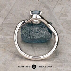 The "Minna" ring in 14K white gold featuring a 1.07-carat Montana sapphire sits on a rough blue stone atop a textured gray surface, with the "Earth's Treasury" logo displayed below.