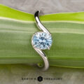 The "Minna" ring in 14K white gold with a 1.07-carat Montana sapphire is displayed on a green, striped leaf. The curved, modern design and the "Earth’s Treasury" logo are visible below.