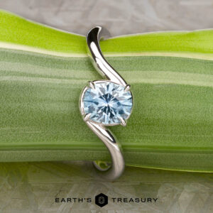 The "Minna" ring in 14K white gold with a 1.07-carat Montana sapphire is displayed on a green, striped leaf. The curved, modern design and the "Earth’s Treasury" logo are visible below.