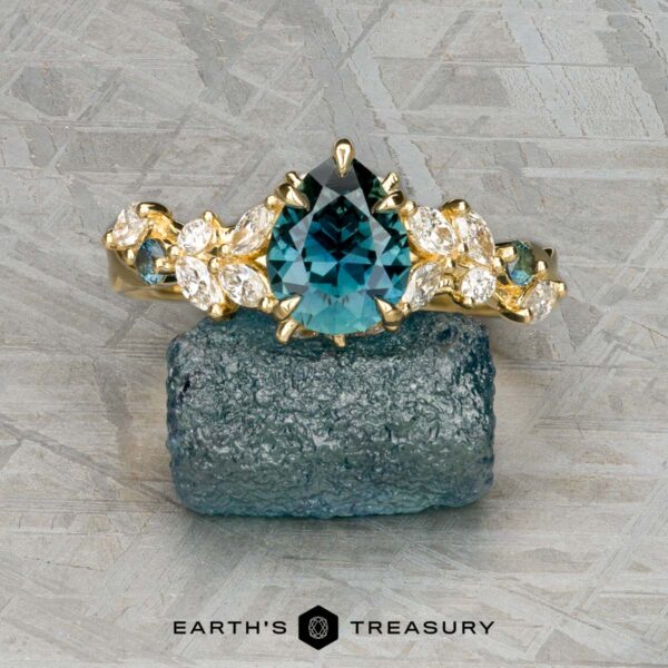 The "Posey" ring in 18K yellow gold with 1.42-carat Montana sapphire The "Posey" ring in 18K yellow gold with a 1.42-carat Montana sapphire is displayed on a rough blue crystal, accented by clear marquise stones. The Earth's Treasury logo appears at the bottom.