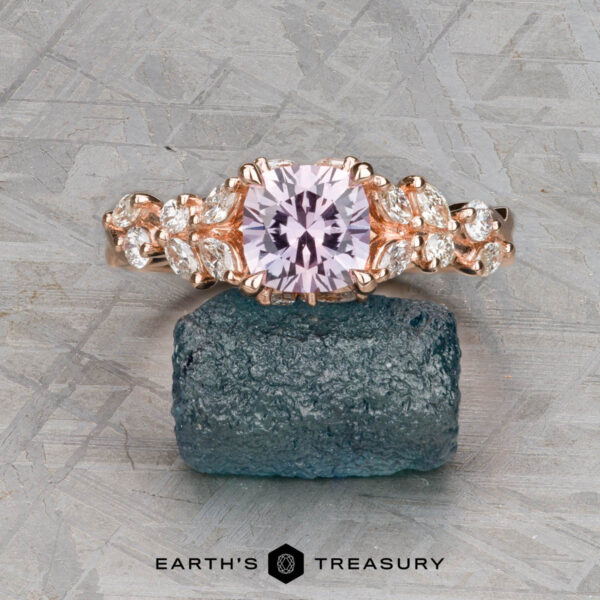 The "Posey" ring in 14K rose gold with 1.38-carat Montana sapphire The "Posey" ring in 14k rose gold, featuring a 1.38-carat Montana sapphire, is showcased on a rough blue-green stone with the "Earth's Treasury" logo beneath.