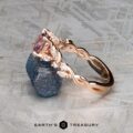 The "Posey" ring in 14k rose gold, set with a 1.38-carat Montana sapphire and marquise-cut diamonds, is displayed on a rough blue stone against a textured gray background.