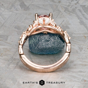 The "Posey" ring in 14k rose gold with a 1.38-carat Montana sapphire is displayed on a blue mineral and patterned surface.