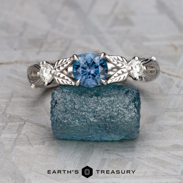 The "Briar" ring in 14K white gold with 0.84-carat Montana sapphire The "Briar" ring in 14K white gold featuring a 0.84-carat Montana sapphire and diamond accents is displayed front-facing on a rough blue crystal, with the "EARTH'S TREASURY" logo at the bottom.