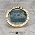 The "Posey" band in 18K yellow gold with a geometric design is displayed upright before a rough blue gemstone, set against a textured gray background with the Earth's Treasury logo below.