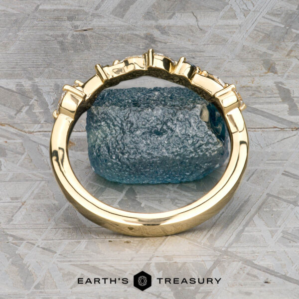 The "Posey" band in 18K yellow gold The "Posey" band in 18K yellow gold with a geometric design is displayed upright before a rough blue gemstone, set against a textured gray background with the Earth's Treasury logo below.