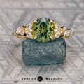 The "Holly" ring in 14K yellow gold, featuring a 2.02-carat Australian sapphire and diamonds, is displayed on a rough blue-green stone with Earth's Treasury branding below.
