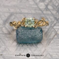 The "Ivey" ring in 14K yellow gold, featuring a leaf design and a 0.85-carat square-cut Montana sapphire, is displayed on rough blue stone with the "EARTH'S TREASURY" logo below.