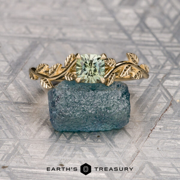 The "Ivey" ring in 14K yellow gold with 0.85-carat Montana sapphire The "Ivey" ring in 14K yellow gold, featuring a leaf design and a 0.85-carat square-cut Montana sapphire, is displayed on rough blue stone with the "EARTH'S TREASURY" logo below.