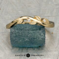The "Ivey" band in 18K yellow gold features a twisted design with delicate leaf accents and two sparkling diamonds, shown resting on a rough blue crystal.
