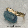 The "Ivey" band in 18K yellow gold features a twisted design with delicate leaf accents and two sparkling diamonds, shown resting on a rough blue crystal.