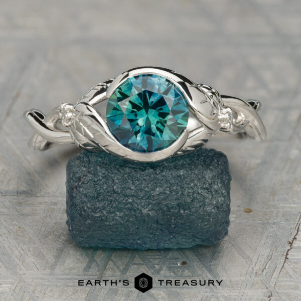 The "Linden" ring in plaitnum with 1.64-carat Montana sapphire The "Linden" ring in platinum features an intricate band and a 1.64-carat round blue-green Montana sapphire, shown upright on a rough blue stone. The text "EARTH'S TREASURY" appears below.