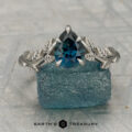 The "Ivey" ring in platinum, featuring a 1.12-carat Montana sapphire and leaf design, is displayed on textured blue stone with "EARTH'S TREASURY" text at the bottom.