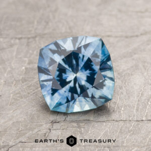 A 1.47-carat aqua blue Montana Sapphire (heated), square-cut with intricate facets, rests on a textured gray surface. The "Earth's Treasury" logo and text appear at the bottom of the image.