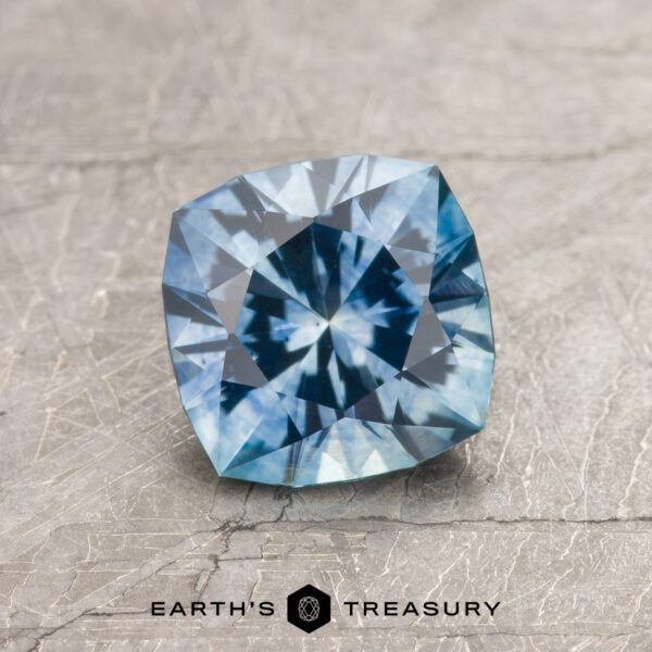 1.47-Carat Aqua Blue Montana Sapphire (Heated) A 1.47-carat aqua blue Montana Sapphire (heated), square-cut with intricate facets, rests on a textured gray surface. The "Earth's Treasury" logo and text appear at the bottom of the image.