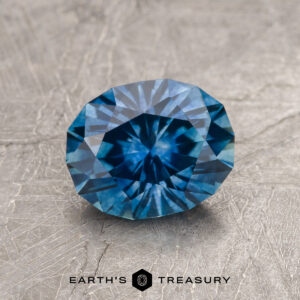 A 1.74-carat medium blue Montana Sapphire (heated) with a geometric cut is displayed on a textured gray surface. The "EARTH'S TREASURY" logo and text are shown at the bottom of the image.