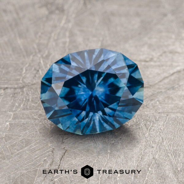 A 1.74-carat medium blue Montana Sapphire (heated) with a geometric cut is displayed on a textured gray surface. The "EARTH'S TREASURY" logo and text are shown at the bottom of the image.