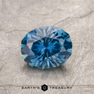 A 1.28-carat medium blue Montana Sapphire (heated), round brilliant-cut with geometric facets, is displayed on a textured gray surface with the "Earth's Treasury" logo below.