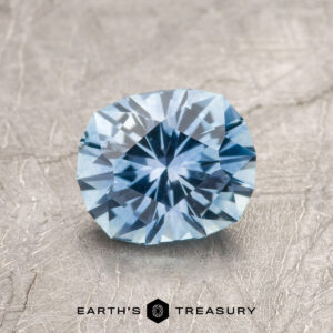 A 1.42-carat sky blue Montana sapphire (heated) rests on a textured surface, with the "Earth's Treasury" logo and text displayed at the bottom.