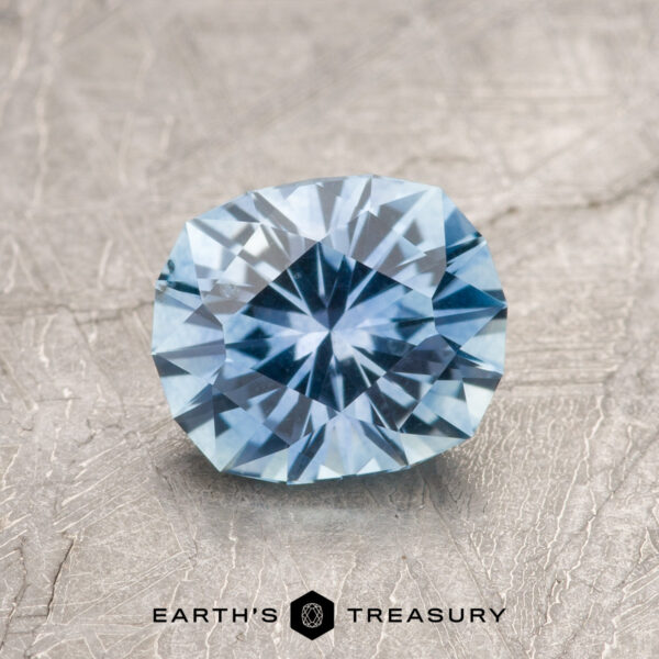 A 1.42-carat sky blue Montana sapphire (heated) rests on a textured surface, with the "Earth's Treasury" logo and text displayed at the bottom.