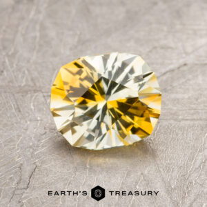 A 1.40-carat Yellow-Gold Particolored Montana Sapphire (Heated) with intricate faceting rests on a textured surface, featuring the Earth's Treasury logo and name at the bottom.