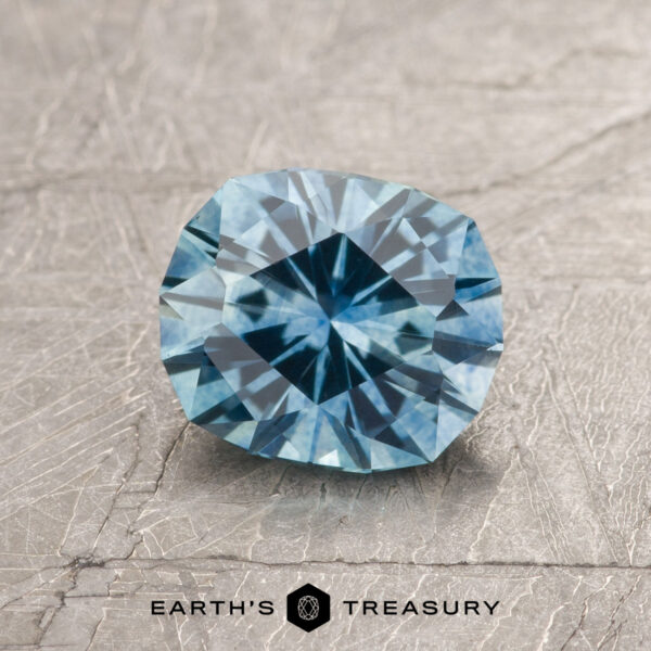 A 1.35-carat Aqua Blue Montana Sapphire (heated, cushion-cut) is shown on a textured gray surface with "EARTH'S TREASURY" and logo at the bottom.